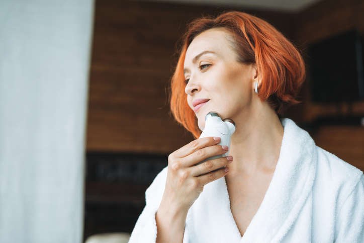 Beauty portrait of smiling woman 35 year in white bathrobe with clean fresh face and hands with red hair doing fasial massage with microcurrent facial massager at bath room, treat yourself, home body care