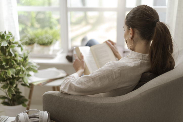 Young woman relaxing at home next to a window, she is sitting on the armchair and reading a book