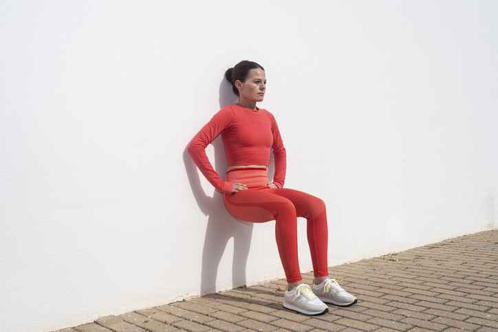 Sporty woman doing squats against a white wall outside in the sun.