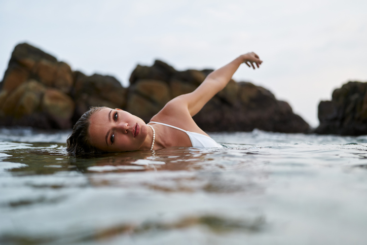 Woman swims gracefully in tranquil lagoon wearing waterproof cosmetics during golden hour. Fashionable swimwear, serene nature backdrop ensures leisure. Female enjoys secluded dip in serene waters.