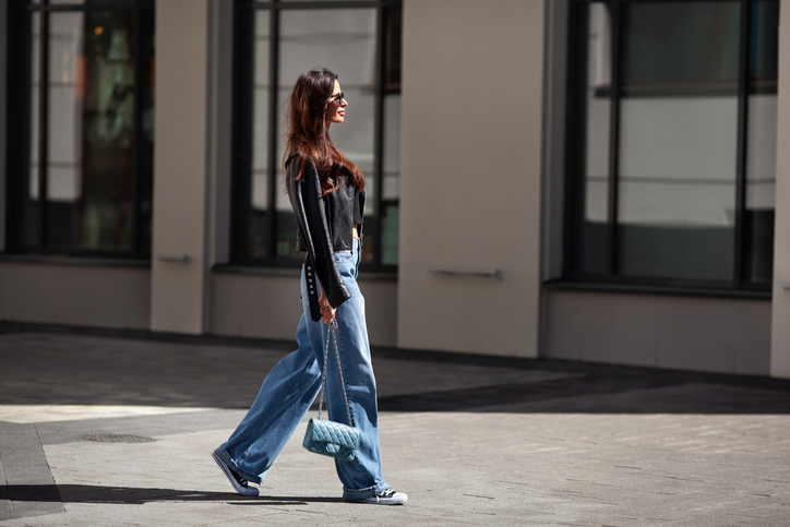 Full length beautiful young brunette woman Wearing black leather jacket and wide blue jeans, sneakers, sunglasses and small handbag with chain, walking street on sunny day
