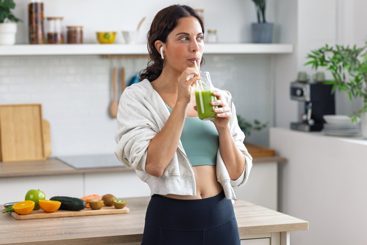 Kind sporty woman drinking a healthy green smoothie standing in the kitchen at home.